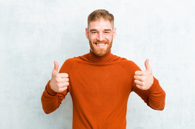 Photo young red head man wearing turtle neck smiling broadly looking happy, positive, confident and successful, with both thumbs up over cement wall