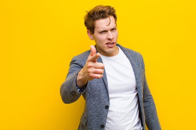 Young red head man with an angry aggressive expression looking like a furious, crazy boss against orange wall