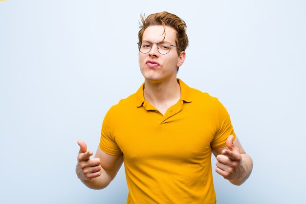 Young red head man with a bad attitude looking proud and aggressive, pointing upwards or making fun sign with hands against blue wall