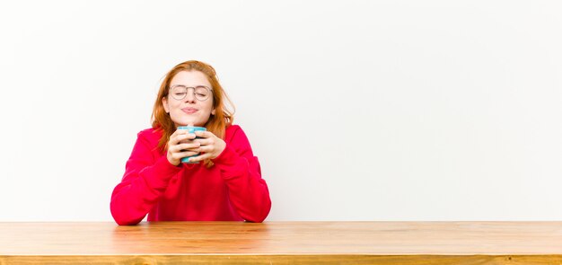 Donna graziosa della giovane testa rossa davanti ad una tavola di legno con una tazza di caffè