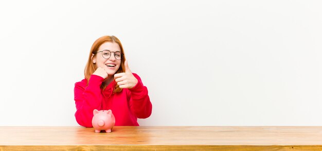 Young red head pretty woman in front of a wooden table with a piggy bank