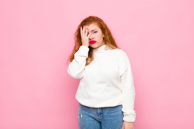 Young red head woman feeling bored, frustrated and sleepy after a tiresome, dull and tedious task, holding face with hand over pink wall