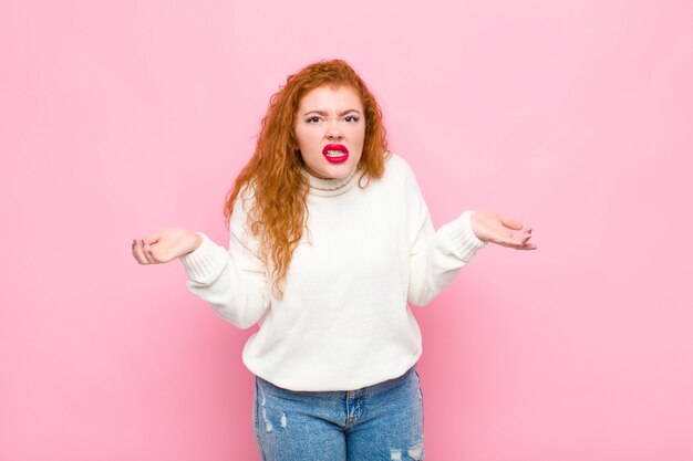 Young red head woman feeling clueless and confused, not sure which choice or option to pick, wondering against pink wall