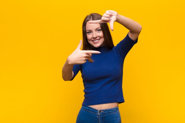 Giovane donna capa rossa sentirsi felice, amichevole e positivo, sorridendo e facendo un ritratto o una cornice con le mani contro la parete piatta