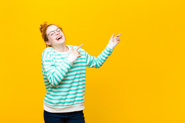 Young red head woman feeling joyful and surprised, smiling with a shocked expression and pointing to the side against orange wall