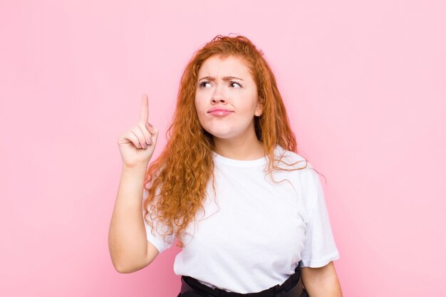 Фото Молодая рыжеволосая женщина, чувствуя себя гением, гордо держащим палец в воздухе после воплощения отличной идеи, произнося эврику на розовой стене