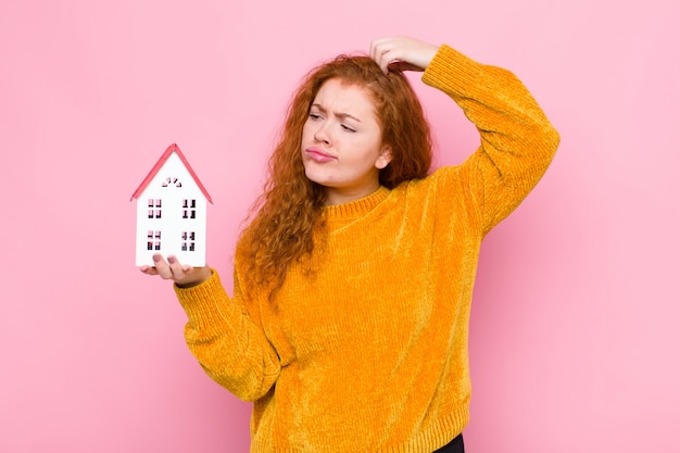 Young red head woman feeling puzzled and confused, scratching head and looking to the side with a house model