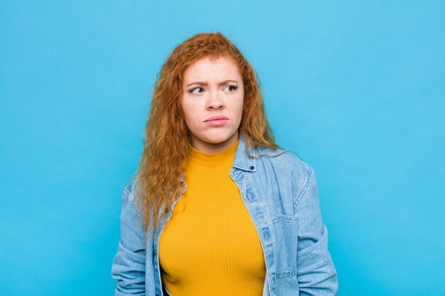 Young red head woman feeling sad, upset or angry and looking to the side with a negative attitude, frowning in disagreement against blue wall