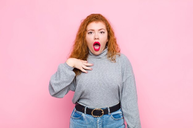 Young red head woman feeling shocked, astonished and surprised, with hand on chest and open mouth, saying who, me? against pink wall