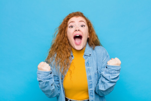 Young red head woman feeling shocked, excited and happy, laughing and celebrating success, saying wow! over blue wall