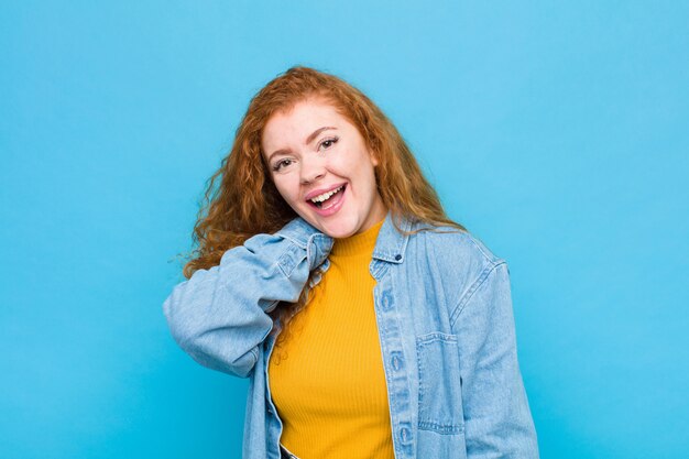 Молодая рыжеволосая женщина весело и уверенно смеется с непринужденной, счастливой, дружеской улыбкой на фоне голубой стены
