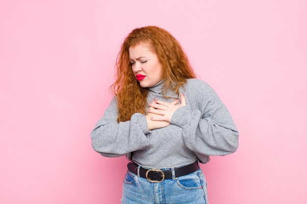 Young red head woman looking sad, hurt and heartbroken, holding both hands close to heart, crying and feeling depressed against pink wall