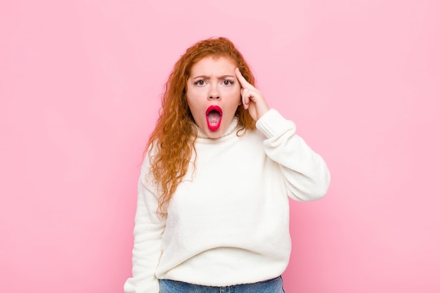 Young red head woman looking surprised, open-mouthed, shocked, realizing a new thought, idea or concept against pink wall