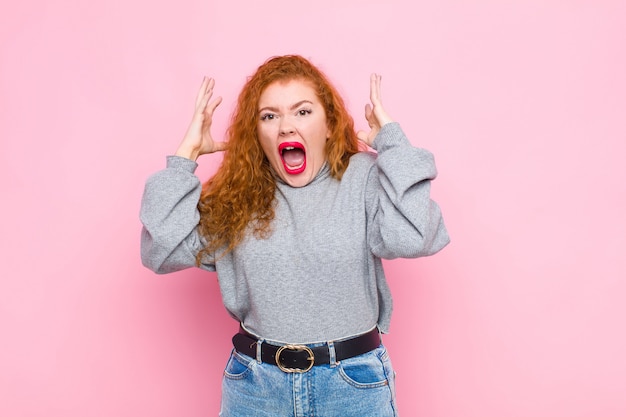 Young red head woman screaming with hands up in the air, feeling furious, frustrated, stressed and upset against pink wall