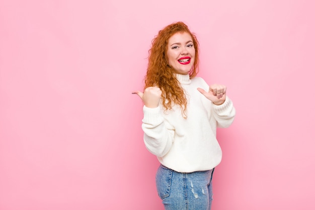 Фото Молодая рыжая женщина весело и небрежно улыбается, указывая, чтобы скопировать пространство сбоку, чувствуя себя счастливой и удовлетворенной у розовой стены