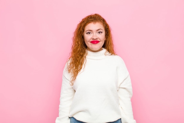 Young red head woman smiling positively and confidently, looking satisfied, friendly and happy against pink wall