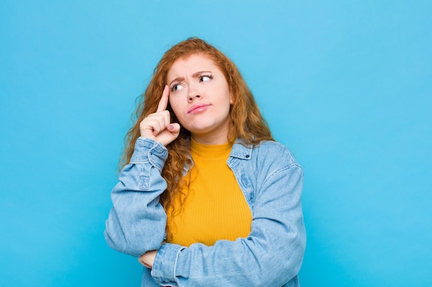 Young red head woman with a concentrated look, wondering with a doubtful expression, looking up and to the side on blue wall