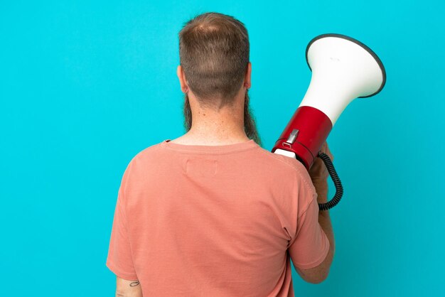 Young reddish caucasian man isolated on blue background holding a megaphone and in back position