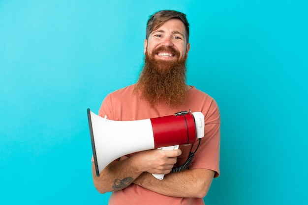 Young reddish caucasian man isolated on blue background holding a megaphone and smiling