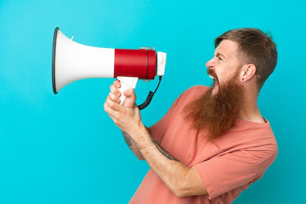 Young reddish caucasian man isolated on blue background shouting through a megaphone to announce something in lateral position