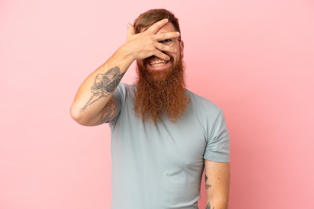 Young reddish caucasian man isolated on pink background covering eyes by hands and smiling