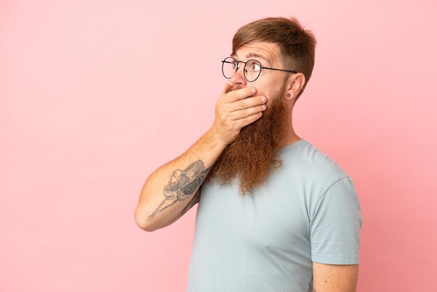 Young reddish caucasian man isolated on pink background doing surprise gesture while looking to the side