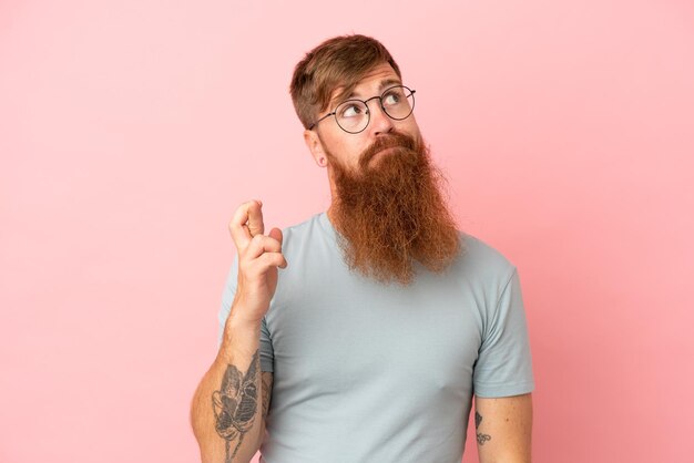 Young reddish caucasian man isolated on pink background with fingers crossing and wishing the best