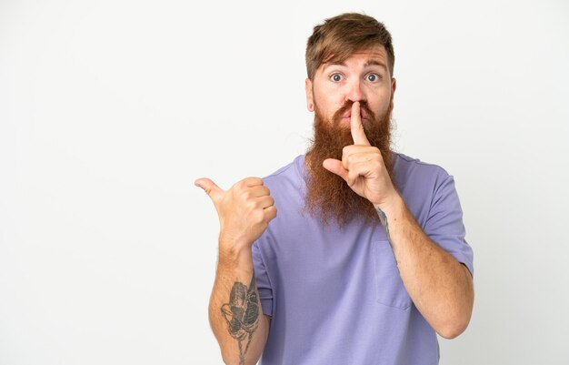 Young reddish caucasian man isolated on white background pointing to the side and doing silence gesture