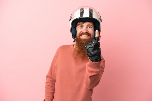 Молодой красноватый кавказец в мотоциклетном шлеме на розовом фоне делает приближающийся жест