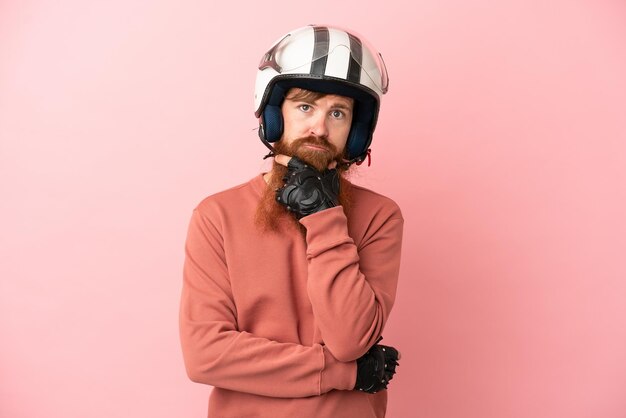 ピンクの背景思考に分離されたオートバイのヘルメットを持つ若い赤みを帯びた白人男性