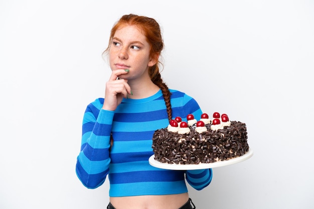 Young reddish woman holding birthday cake isolated on white background having doubts and thinking