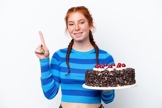 Молодая красноватая женщина держит торт ко дню рождения на белом фоне, показывая и поднимая палец в знак лучшего