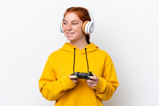 Young reddish woman playing with a video game controller isolated on white background looking to the side and smiling