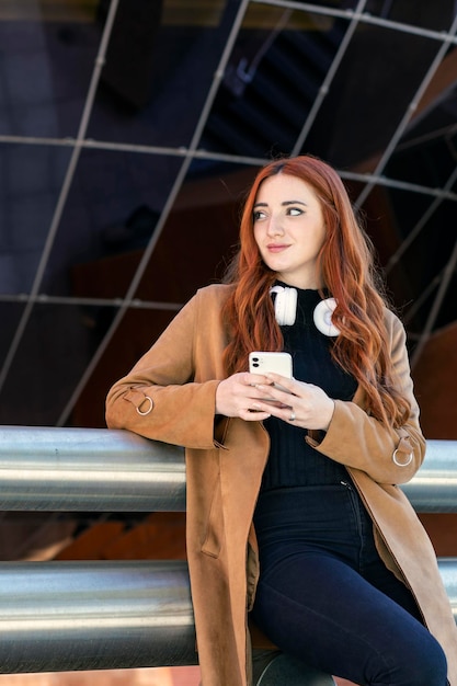 Young redhaired girl looking to the side with her cell phone in her hand and wireless headphones listening to music on a city street