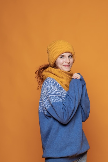 Young redhaired girl in a warm hat and a knitted sweater on a yellow background