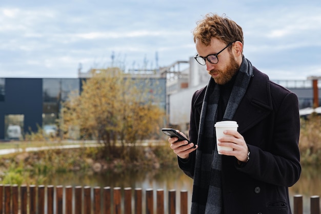 도시 스타일을 배경으로 안경을 쓴 젊은 빨간 머리 남자는 스마트폰으로 메일을 확인하고 메신저로 통신하고 커피를 마신다 온라인 커뮤니케이션