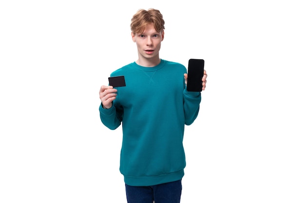 Photo young redhaired man with a blue sweater shows a plastic card mockup and a smartphone on a white