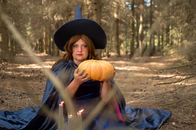 A young redhaired witch in a cloak and a pointed hat with a pumpkin in her hands in the forest