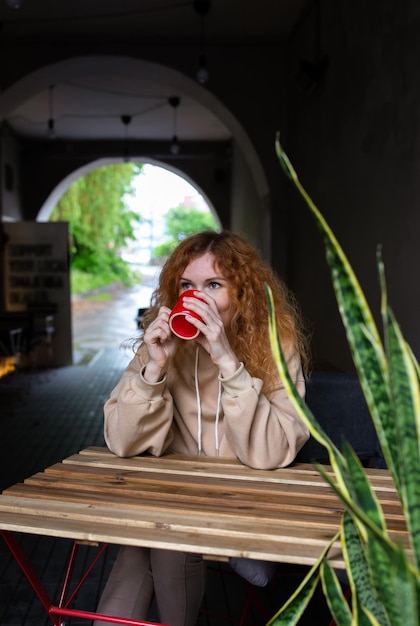 Young redhaired woman alone drinking a drink in a terrace cafe\
on a rainy day