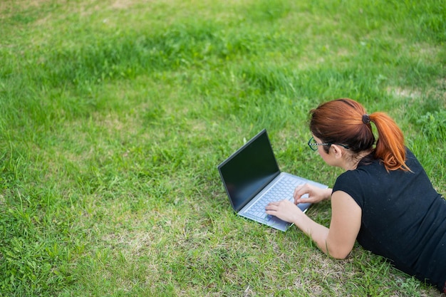 Young redhaired woman remotely works out lying on green grass Freelancer girl prints on a laptop outdoors