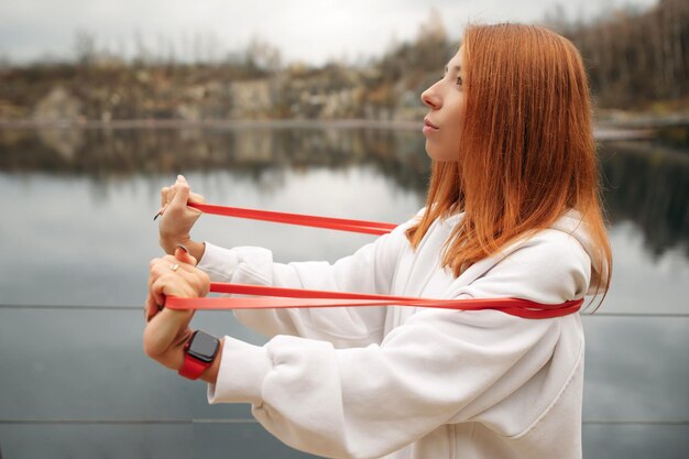 Photo young redhaired woman in a white hoodie doing streching with resistance band in the park near the lake healthy and sport concept