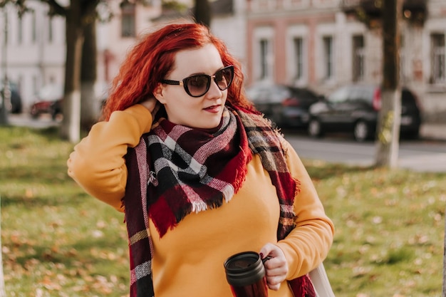 Young redhaired woman in yellow sweater and sunglasses holds thermo mug with coffee in her hands Lady walks through city and park on sunny autumn day