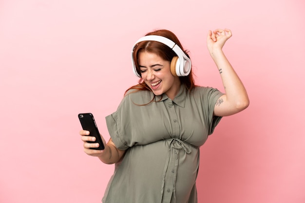 Young redhead caucasian woman isolated on pink background pregnant and dancing while listening music