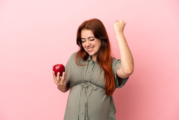 Young redhead caucasian woman isolated on pink background pregnant and holding an apple while celebrate a victory