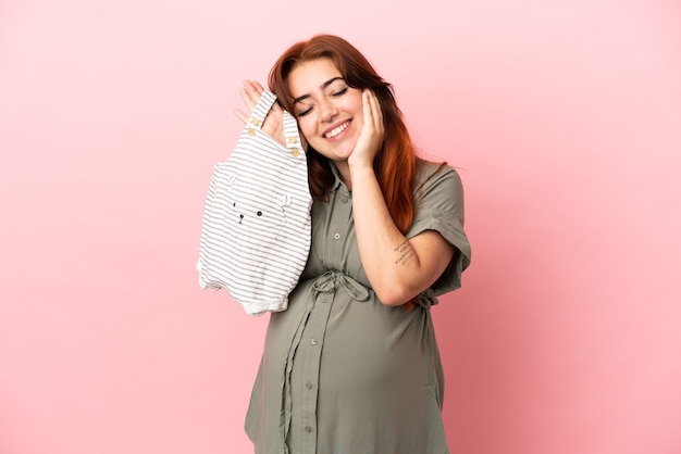 Young redhead caucasian woman isolated on pink background pregnant and holding baby clothes