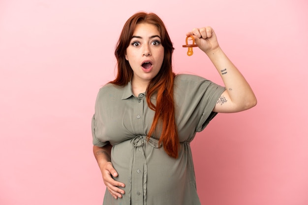 Young redhead caucasian woman isolated on pink background pregnant and holding a pacifier with surprised expression