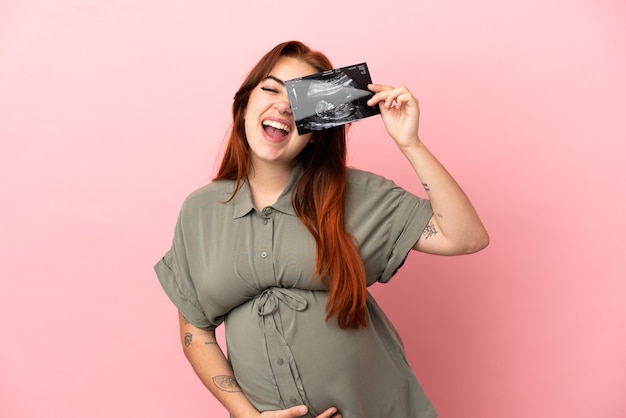 Young redhead caucasian woman isolated on pink background pregnant and holding an ultrasound