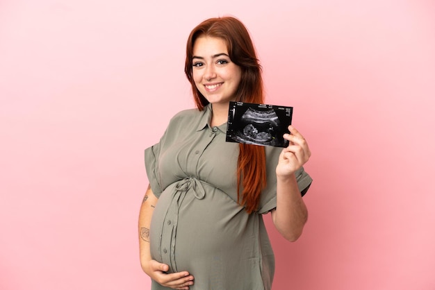 Young redhead caucasian woman isolated on pink background pregnant and holding an ultrasound