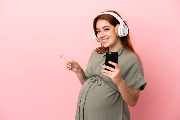 분홍색 배경에 고립 된 젊은 빨간 머리 백인 여성 임신 및 측면을 가리키는 동안 음악을 듣고