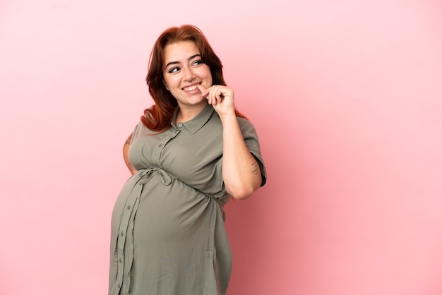 Young redhead caucasian woman isolated on pink background pregnant and thinking